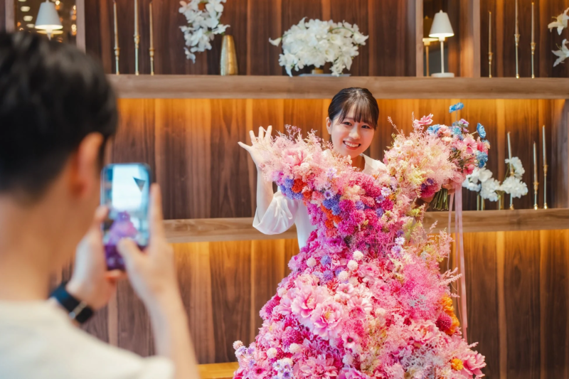 花で飾られたドレスのような衣装をまとった女性が笑顔でポーズをとり、別の人物がスマートフォンで写真を撮っている様子。華やかで楽しい雰囲気が伝わります。