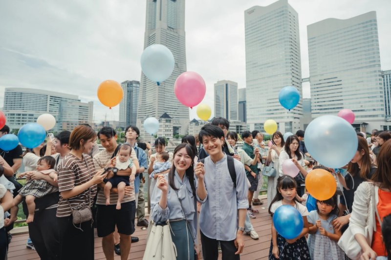 多くの人々がカラフルな風船を持ち、笑顔で集まっている屋外イベントの様子。横浜の都市景観を背景に、家族連れや友人同士が楽しんでいる賑やかな光景です。