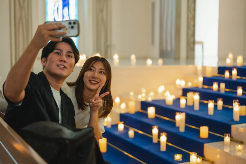 青いカーペットの階段に並べられた無数のキャンドルの光を背景に、若いカップルがスマートフォンで自撮りを楽しんでいる様子。ロマンチックな雰囲気が漂うイベント会場やチャペルのような場所で、笑顔を見せている。