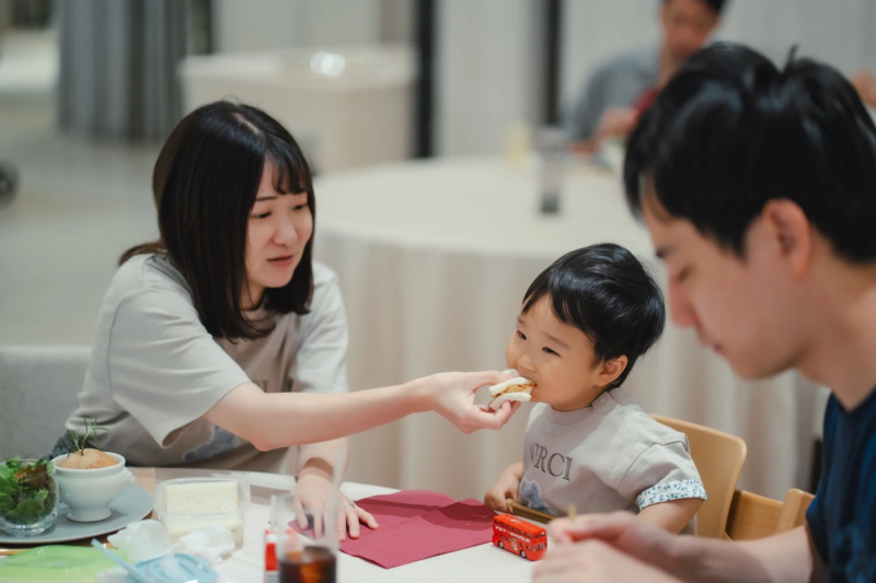 女性が子供にサンドイッチを食べさせている、和やかな家族の食事風景です。男性も同席しており、テーブルには他の料理や小さなバスのおもちゃが見えます。