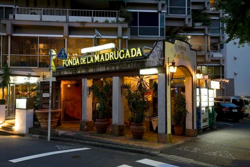 「FONDA DE LA MADRUGADA」という看板が明るく輝く建物の夜の外観です。アーチ状の入口と柱、植木が特徴的で、異国情緒あふれるレストランの雰囲気が伝わります。