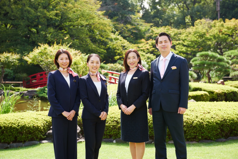 日本庭園を背景に、ビジネススーツと制服を着用した4人のプロフェッショナルが笑顔で並んで立っています