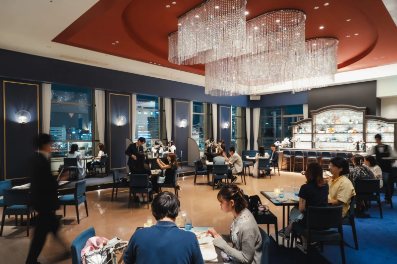 豪華なシャンデリアが輝くレストランバーの夜景。人々が食事や会話を楽しんでおり、洗練された内装が特徴の都会的な空間です。