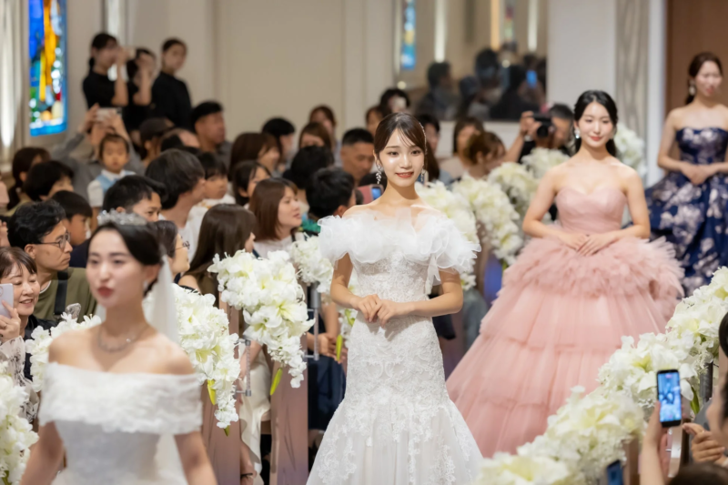 複数の女性がウェディングドレスやカラードレスを着用し、観客が見守る中でランウェイを歩いている様子です。ブライダルイベントやファッションショーの一場面と考えられます。