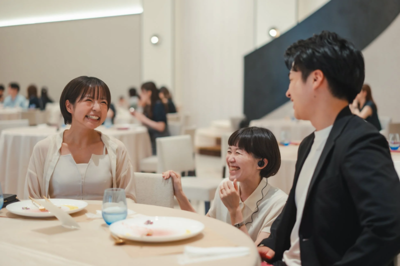明るい室内で、テーブルを囲んで笑顔で会話を楽しむ男女3人の様子。和やかな雰囲気の懇親会やビジネスイベントの一場面と見られます。