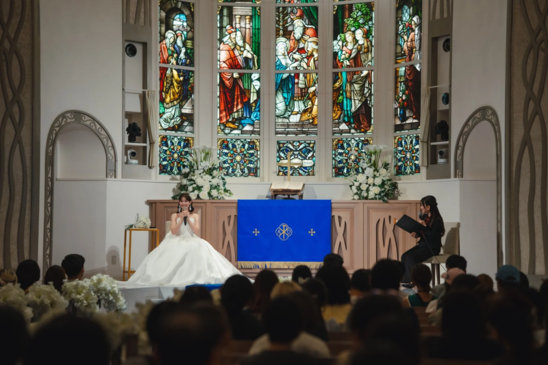 結婚式のチャペルで、白いウェディングドレスを着た花嫁がマイクを手に笑顔で歌っているかスピーチしている様子。背景には美しいステンドグラスがあり、祭壇には青い布がかけられ、手前には参列者が集まっています。