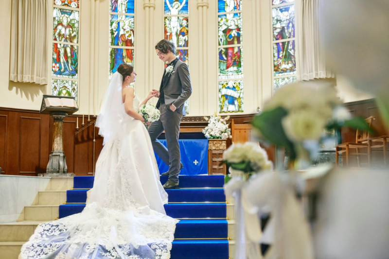 教会で行われている結婚式の様子です。新郎新婦が青い絨毯の階段に立ち、お互いを見つめ合っています。背景には美しいステンドグラスが見えます。