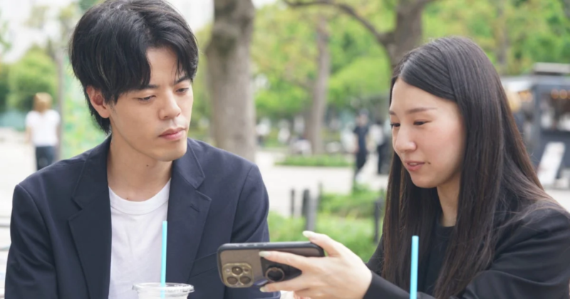 若い男女が屋外のカフェのような場所で、女性が持つスマートフォンを一緒に見ながら会話を楽しんでいます