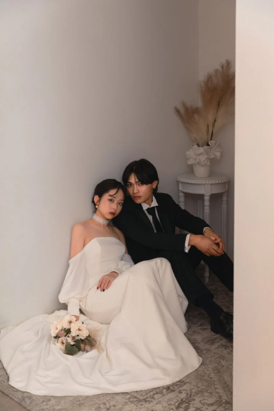 白いウェディングドレス姿の女性と黒いスーツ姿の男性が壁にもたれて床に座っているポートレート。